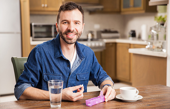 Ein junger Mann sitzt am Küchentisch und hält eine Tablettenbox in der Hand.