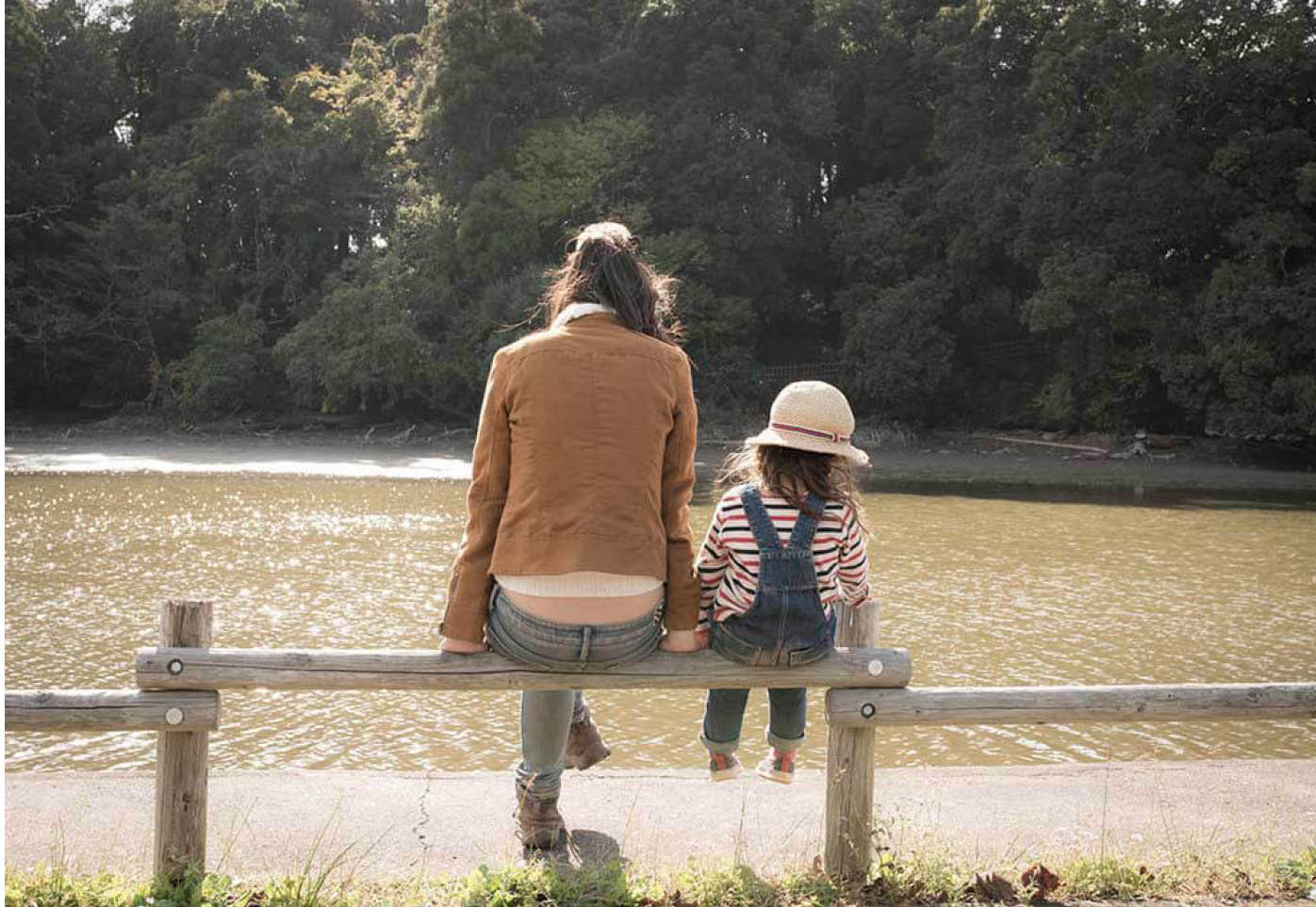 Eine Frau und ein kleines Mädchen sitzen auf einem Holzzaun und blicken auf einen Teich.