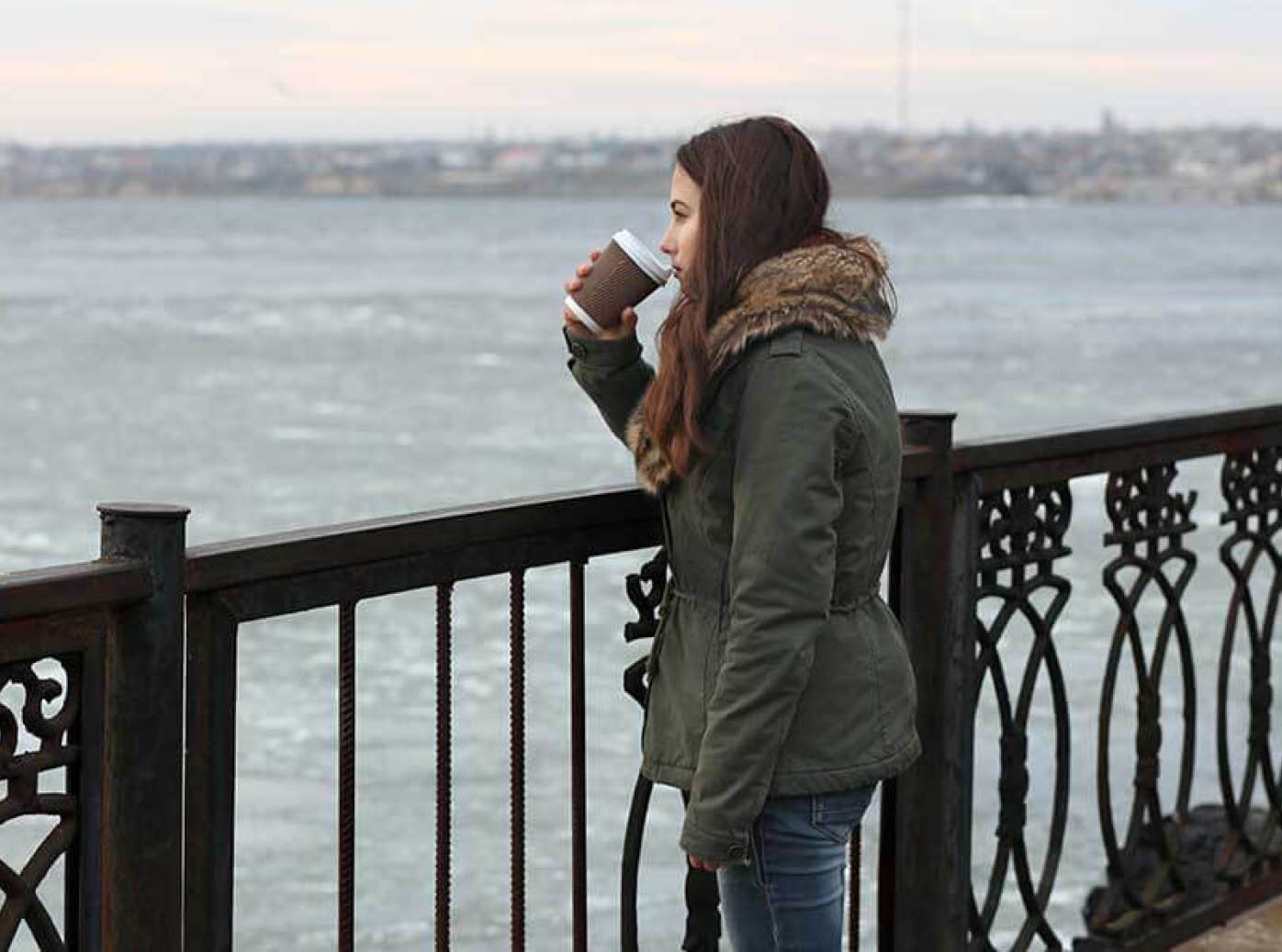 Eine nachdenkliche junge Frau steht mit einem Kaffeebecher in der Hand auf einer Strandpromenade.